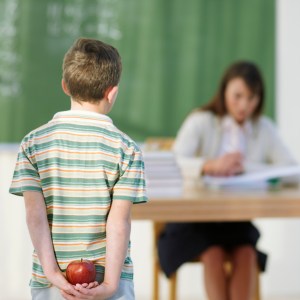 Boy (8-9) holding apple behind back, looking at teacher in classroom, focus on boy